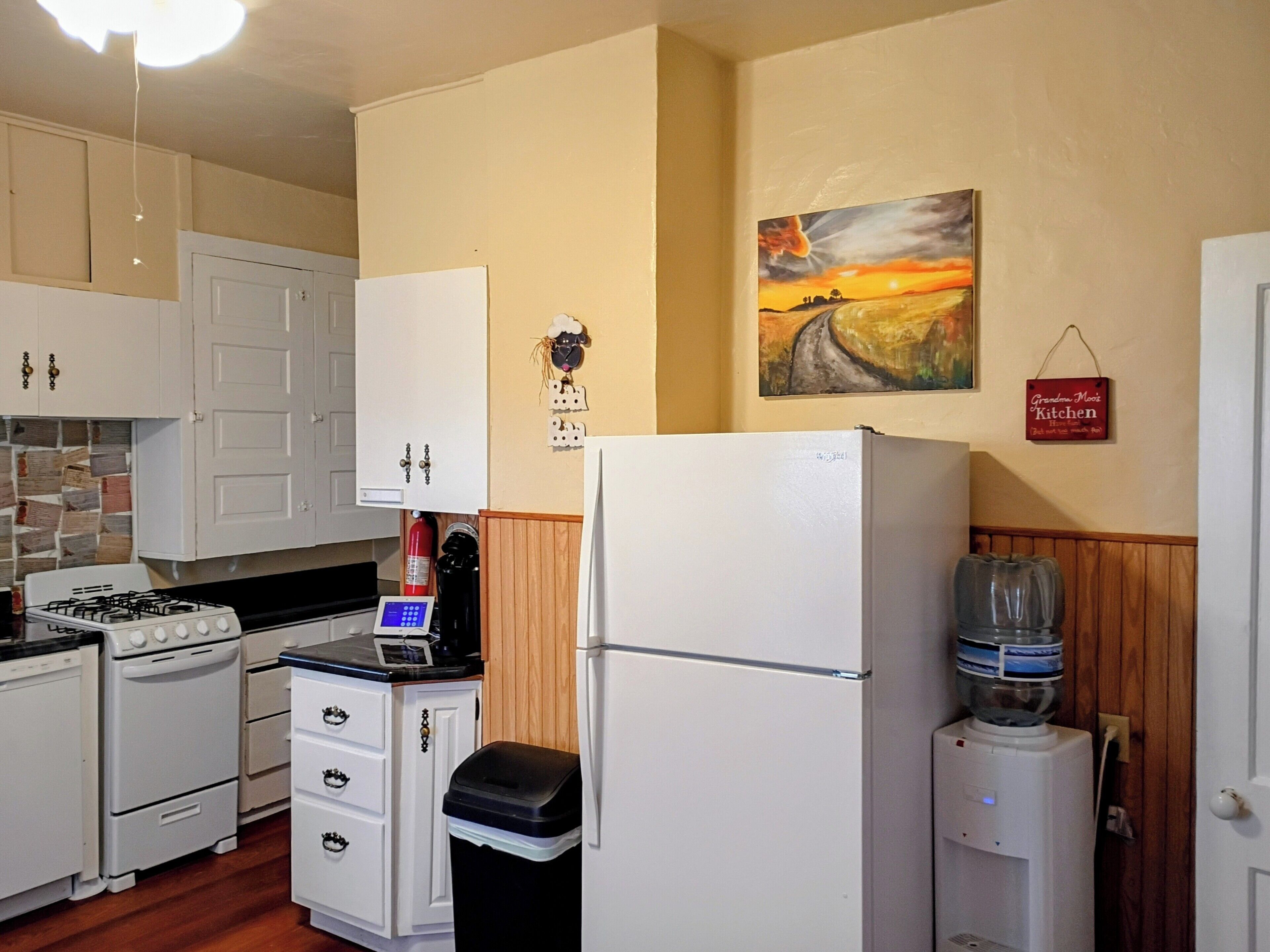 A compact kitchen with white cabinets, a stove, a refrigerator, a water dispenser, and a small trash bin.