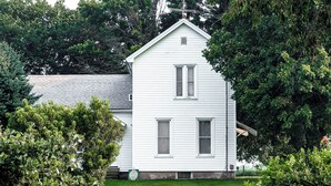 Uma casa branca de dois andares com telhado empena, cercada por vegetação e um quintal murado.