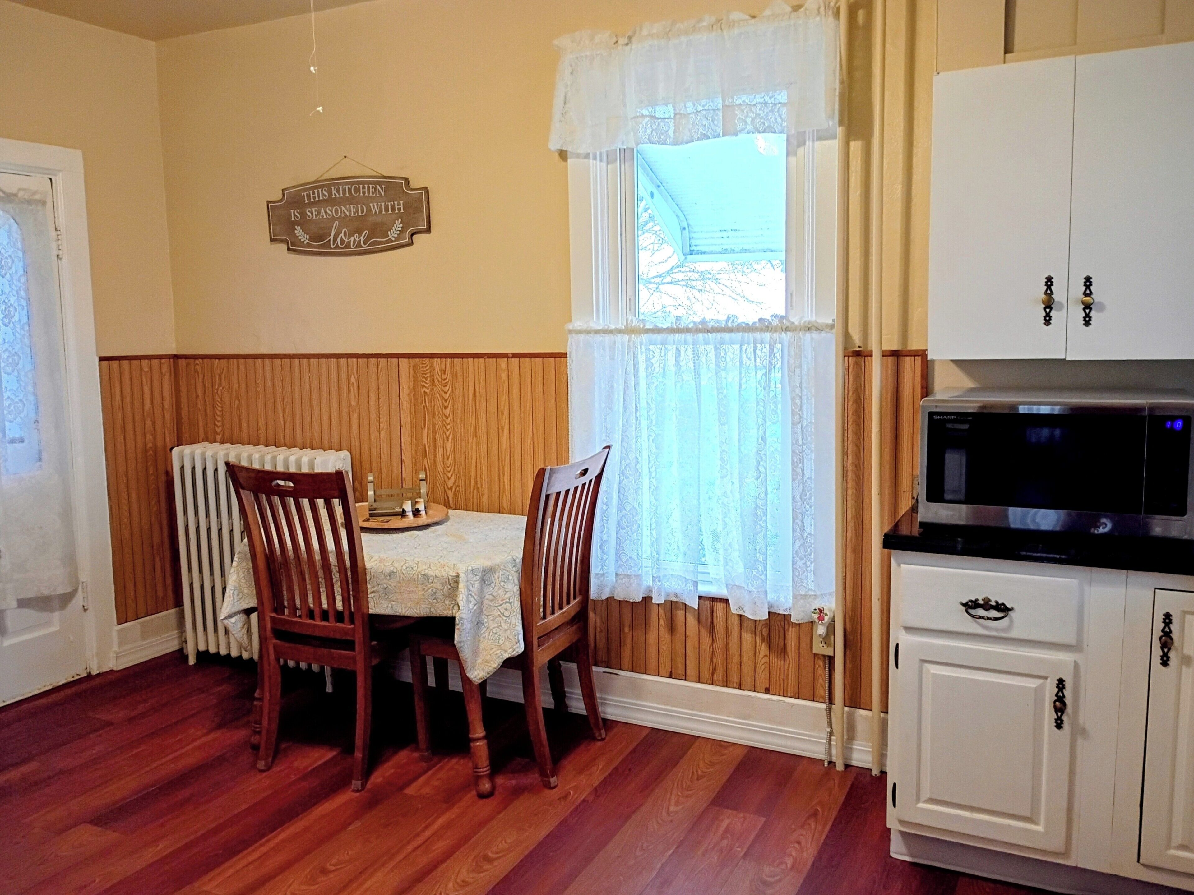 A dining area with a table and chairs, a window with lace curtains, a kitchen with cabinets and a microwave, and a sign on the wall.