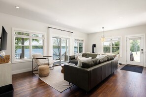A spacious living room with a large sofa, a chair, and a TV. There are large windows offering a view of a lake. - Lakefront Cottage on Lake Leelanau | Dock, Fire Pit, Near M-22 & Dunes (Lake Leelanau)