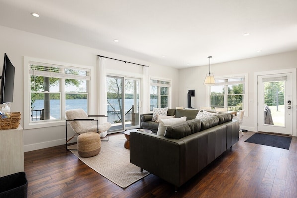 Un salon spacieux avec un grand canapé, un fauteuil et un téléviseur. De grandes fenêtres offrent une vue sur un lac.
