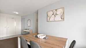 Una habitación moderna con una mesa de comedor de madera, sillas grises, una pared blanca con una obra de arte enmarcada, una mesita de noche y un espejo.