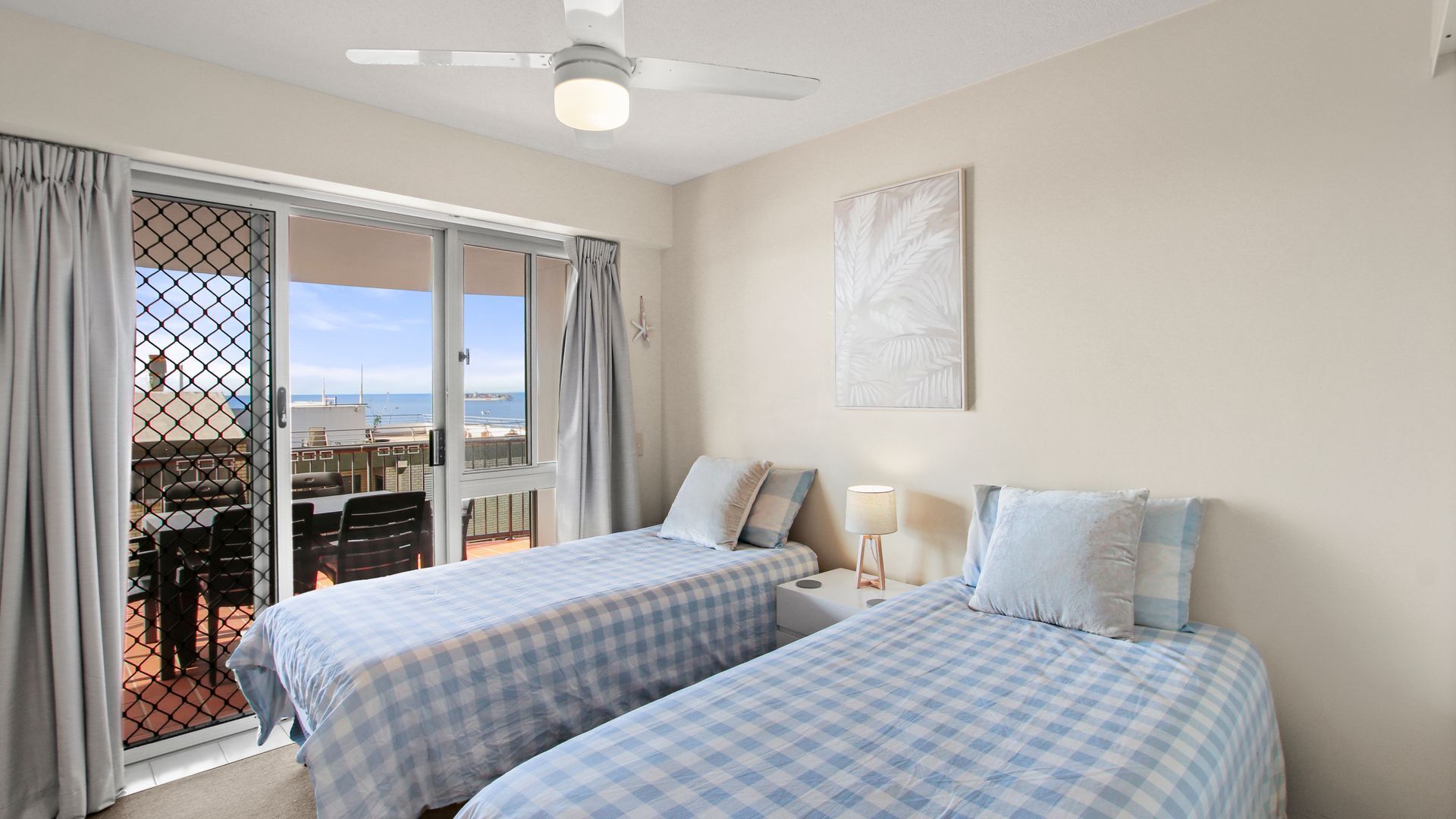 A bedroom with two beds, a ceiling fan, a window with curtains, a table with chairs, and a view of the sea.