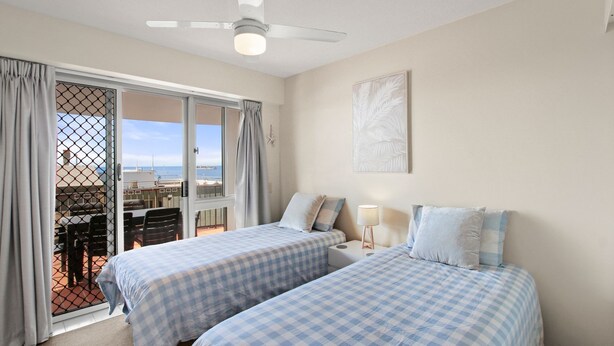 Un dormitorio con dos camas, ventilador de techo, ventana con cortinas, una mesa con sillas y vista al mar.
