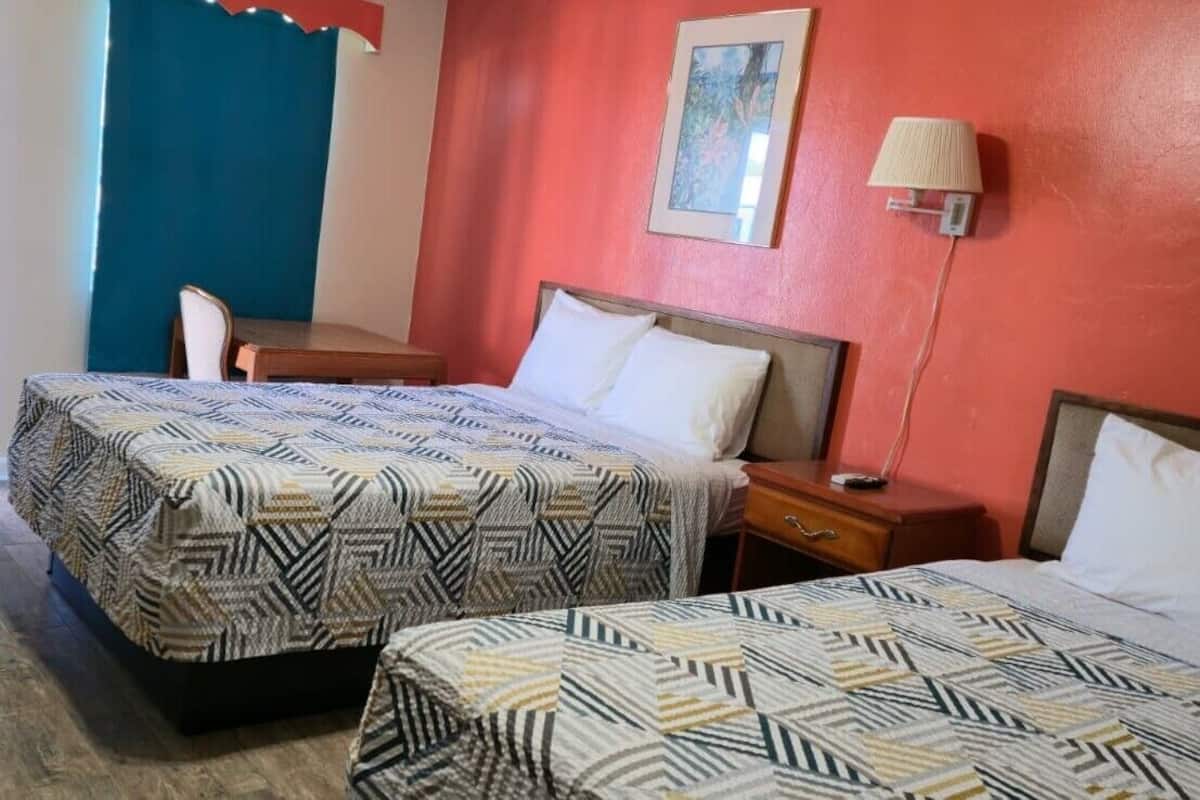 A hotel room with two beds, a desk, a chair, a lamp, and a painting on the wall.