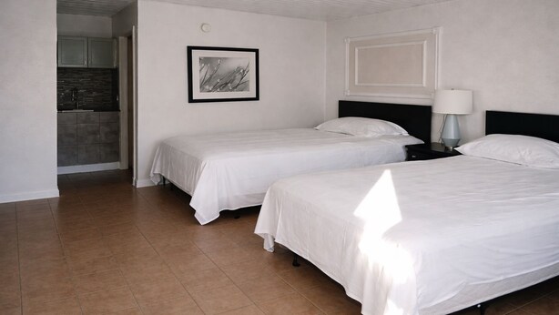 A hotel room with two single beds, a small bedside table with a lamp, a framed picture on the wall, and a kitchenette in the background. - Siesta Motor Inn Kissimmee Near Disney World (Kissimmee)