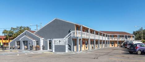A motel with a grey building and a yellow building with balconies. There are cars parked in the lot.