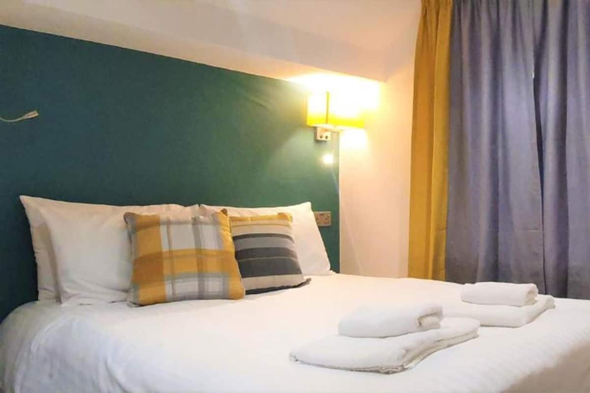 Een netjes opgemaakt bed met witte lakens en twee kussens, een set opgevouwen handdoeken en een wandlamp.