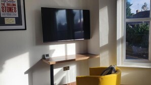A room with a flat-screen TV mounted on the wall, a wooden shelf below, and a yellow chair in front of a window. - Oyo Studio @74 (Swindon)