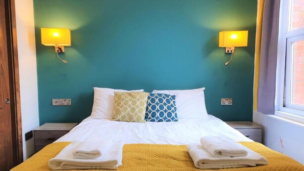A neatly made bed with a yellow quilt, two pillows, and folded towels on a teal-colored wall with two wall-mounted lamps. - Oyo Studio @74 (Swindon)