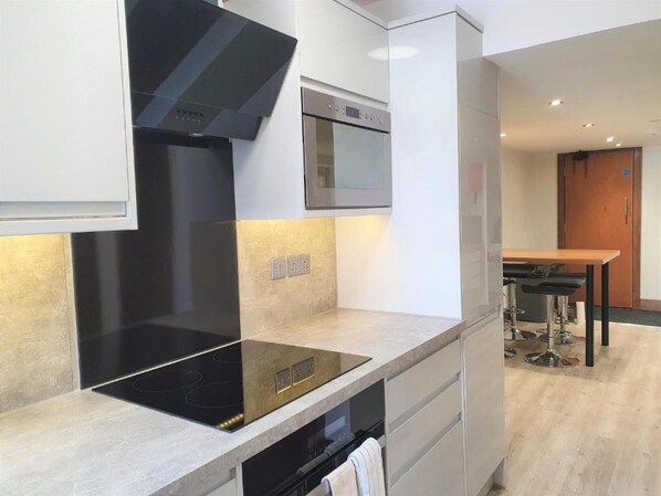 A modern kitchen with a black induction cooktop, a stainless steel oven, and a built-in range hood. There is a wooden dining table with bar stools in the background. - Oyo Studio @74 (Swindon)
