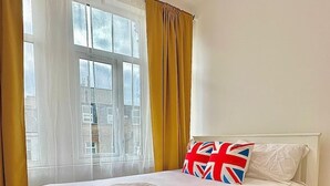 A white bed with a Union Jack pillow, a window with yellow curtains, and a wooden floor. - 10-minute Ride to Central London A (London)