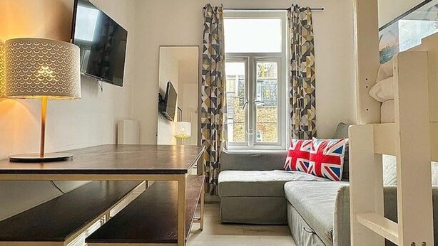 A modern, well-lit room with a desk, a television mounted on the wall, a window with patterned curtains, and a bed with a Union Jack pillow. - 10-minute Ride to Central London A (London)
