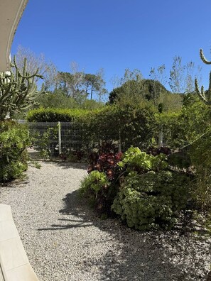 A gravel pathway leading to a fenced garden area with various plants and trees. - Appartement rez-de jardin avec terrasse et parking privé à Calvi à 150m de la plage (CALVI)
