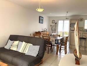 Een gezellige woonkamer met een donkere bank, een houten eettafel en stoelen. Er is een keukenhoek met een fornuis en een hangende decoratie van een zeilboot.