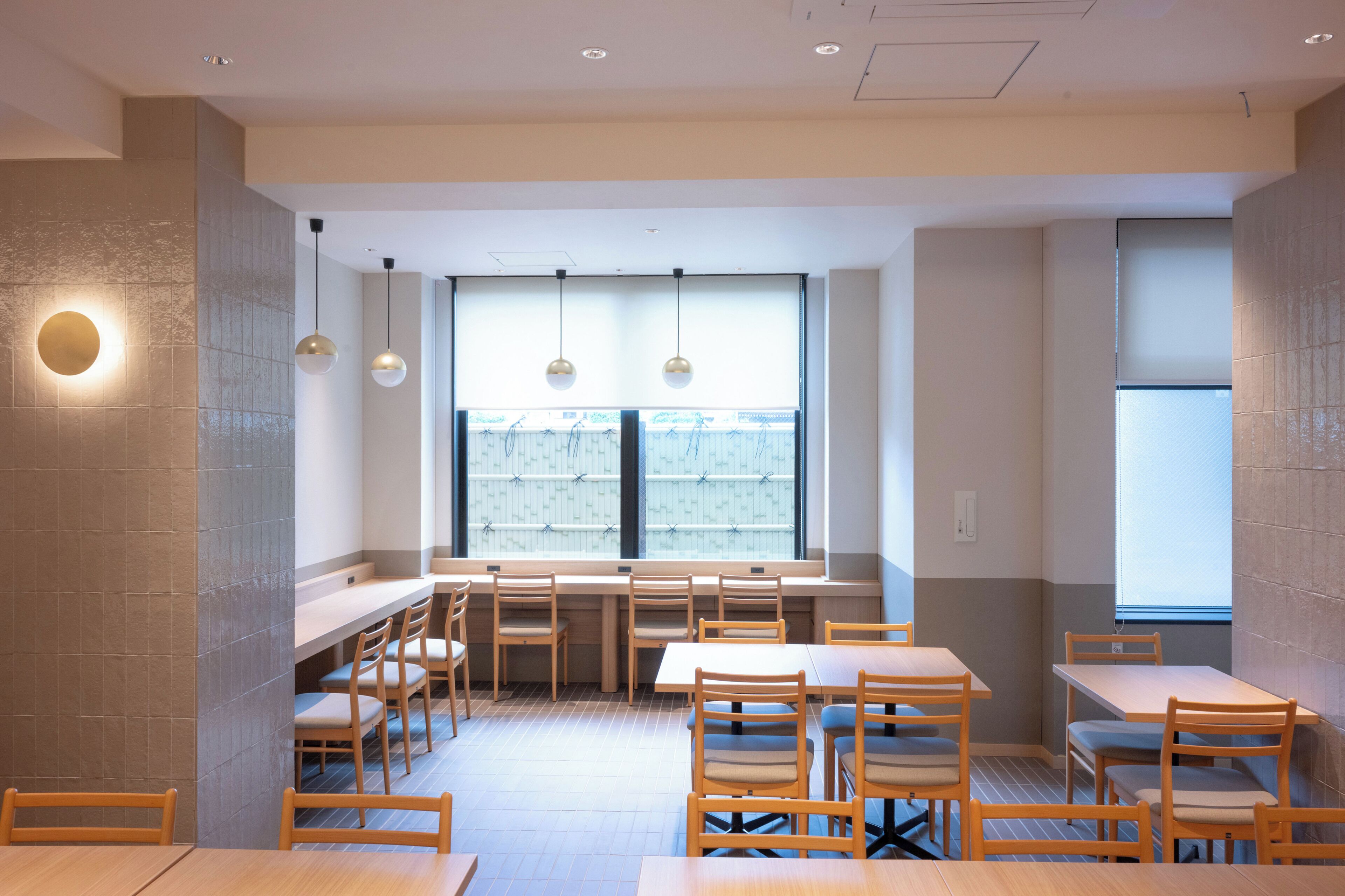 Interior de restaurante com várias mesas e cadeiras, uma grande janela com persianas e luminárias pendentes.