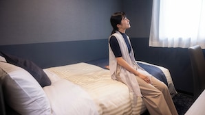 A single bed with a striped blanket and two pillows, a window with sheer curtains, and a plain wall. - Hotel KAN-RAKU Premier Utsunomiya-Eki (Utsunomiya)