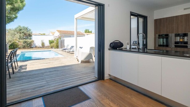 A modern kitchen with a large window, a pool area with lounge chairs, and a view of the surrounding neighborhood. - Ovalie - Stunning Family Home with Swimming Pool Just Steps from the Ocean (RIVEDOUX PLAGE)