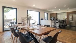 A modern dining area with a wooden table, black chairs, and a kitchen in the background. - Ovalie - Stunning Family Home with Swimming Pool Just Steps from the Ocean (RIVEDOUX PLAGE)