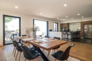 A modern dining area with a wooden table, black chairs, and a kitchen in the background. - Ovalie - Stunning Family Home with Swimming Pool Just Steps from the Ocean (RIVEDOUX PLAGE)