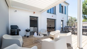 A covered outdoor seating area with white chairs, a grill, and a small table. - Ovalie - Stunning Family Home with Swimming Pool Just Steps from the Ocean (RIVEDOUX PLAGE)