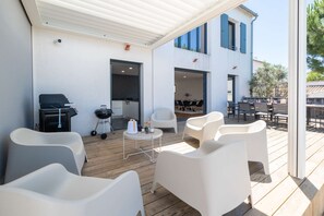 A covered outdoor seating area with white chairs, a grill, and a small table. - Ovalie - Stunning Family Home with Swimming Pool Just Steps from the Ocean (RIVEDOUX PLAGE)