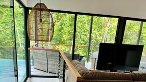 Un interior modern amb una escala, un sofà, una televisió i grans finestres amb vista a vegetació.