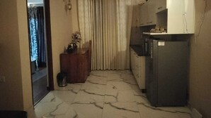 A compact kitchen with a refrigerator, a wooden cabinet, and a curtain. - Luxury 1BHK Retreat in Sector 106 - Pool, Gym & Premium Comfort (Gurugram)