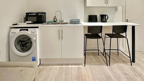 A compact kitchen with a washing machine, microwave, sink, and two black chairs at a white table. - Station-side Top-floor Studio (London)