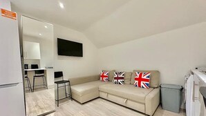 A compact living space with a kitchenette, a sofa with British flag cushions, and a flat-screen TV on the wall. - Station-side Top-floor Studio (London)