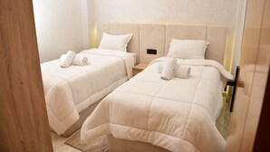 A hotel room with two beds, each with a white duvet and pillows, a wooden nightstand, and a wall-mounted light fixture.