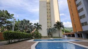 Un hotel con zona de piscina, rodeado de palmeras y un edificio alto al fondo.