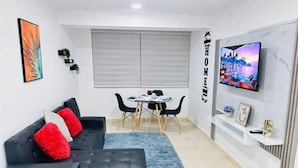 Una sala de estar moderna con un sofá negro, cojines rojos, una mesa de comedor con sillas, un televisor montado en la pared y una ventana con persianas.