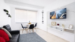 Un salón moderno con un sofá negro, una mesa de comedor con sillas, una alfombra gris, un televisor montado en la pared y una ventana con persianas.