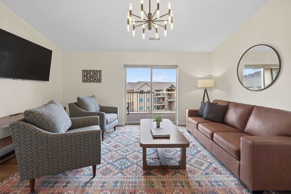 Un salon avec un tapis à motifs, un canapé en cuir marron, des fauteuils à motifs, une table basse avec une plante, un téléviseur à écran plat fixé au mur et une grande fenêtre donnant sur des immeubles.