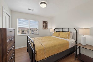 Une chambre à coucher comprenant un lit, des tables de chevet, une commode, une fenêtre avec des stores et un luminaire au plafond.