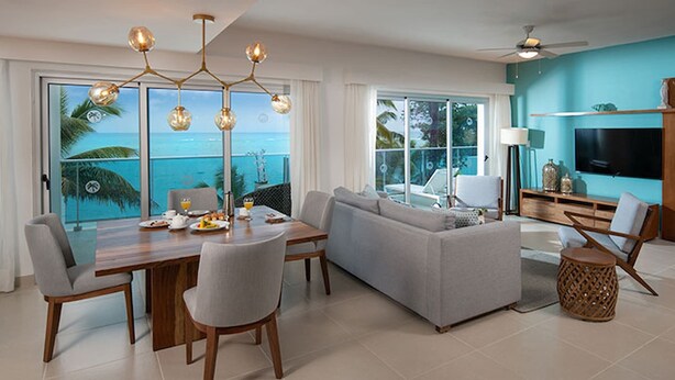 A modern living room with a large sofa, dining table, and chairs. There is a TV mounted on the wall and a ceiling fan. The room has large windows with a view of the ocean. - Imperial Suite 2 Bedroom Ocean View With Kitchen (Sosúa)