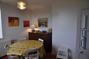 A dining area with a round table covered by a patterned tablecloth, a lamp on a wooden cabinet, and wall art. - La Maison Neuve - COMELLE (LA) (COMELLE (LA))