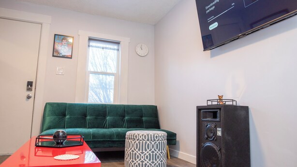 A living room with a green sofa, a red coffee table, a patterned ottoman, a black speaker, and a wall-mounted TV displaying an update package. - Marda Loop Blue Bungalow (Calgary)