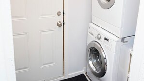 A laundry room with a front-loading washer and dryer, a white door, and a black carpet. - Marda Loop Blue Bungalow (Calgary)