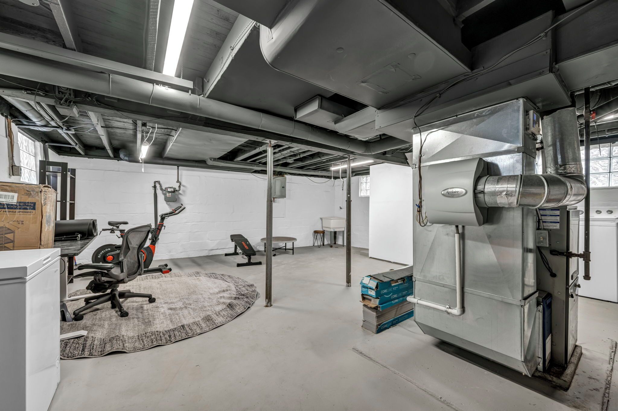 Une pièce spacieuse, aménagée dans un style industriel, avec une grande machine sur la droite, du matériel d’exercice au centre et un mur blanc avec une fenêtre sur la gauche.