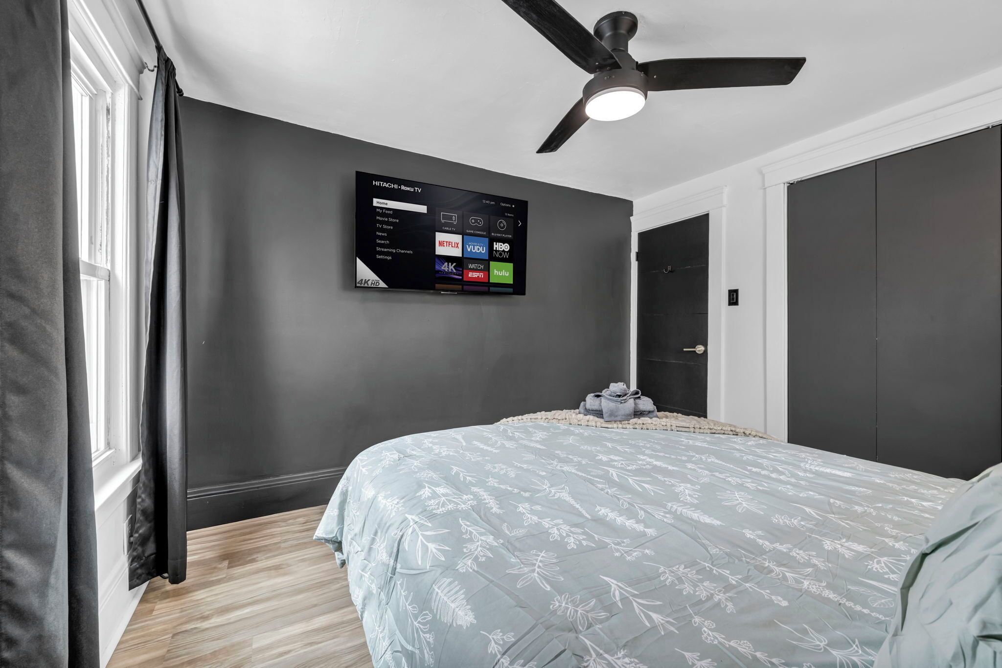 Une chambre à coucher comprenant un lit, un ventilateur de plafond, un téléviseur fixé au mur diffusant des services de streaming et une fenêtre avec des rideaux.