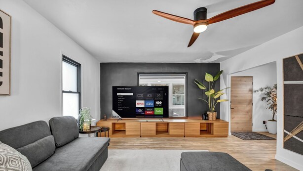 Una sala de estar moderna con un televisor de pantalla plana grande, un mueble de entretenimiento de madera, sofás grises y un ventilador de techo.