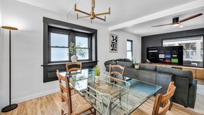 Una sala de estar moderna con una mesa de comedor de vidrio, sofás oscuros y un televisor grande que muestra servicios de streaming.