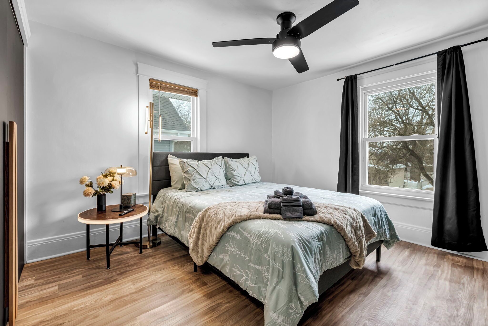 A bedroom with a bed, a ceiling fan, a window with curtains, a small table with a lamp, and a vase of flowers.