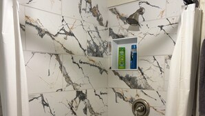 A shower with marble-patterned tiles, a showerhead, and a shelf with toiletries. - Large, well equipped Studio with Queen Bed and new bathroom. (Tucson)