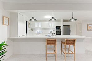 Eine moderne Küche mit weißen Schränken, einer großen Kochinsel mit zwei Barhocker, Pendelleuchten und einem Kühlschrank.