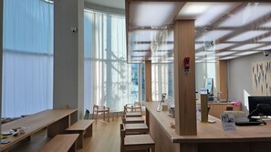 Un espacio de oficina moderno con escritorios y sillas de madera, y un gran ventanal con cortinas translúcidas.
