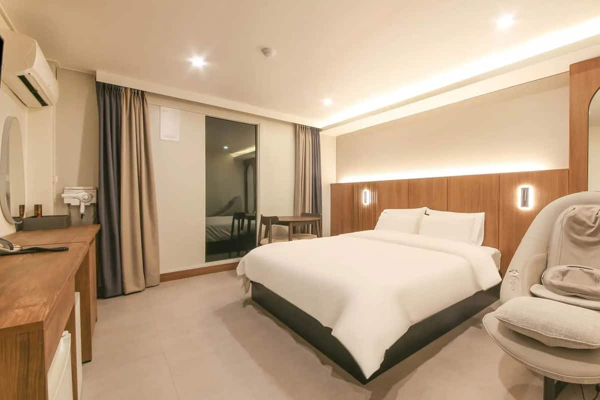 A hotel room with a bed, a desk with a phone, a chair, a mirror, and a wall-mounted air conditioner.