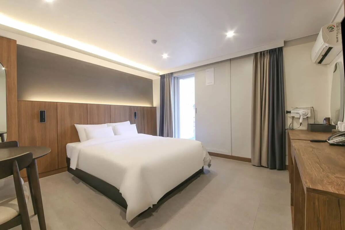 A hotel room with a bed, a desk, a chair, a mirror, a TV, and an air conditioning unit.